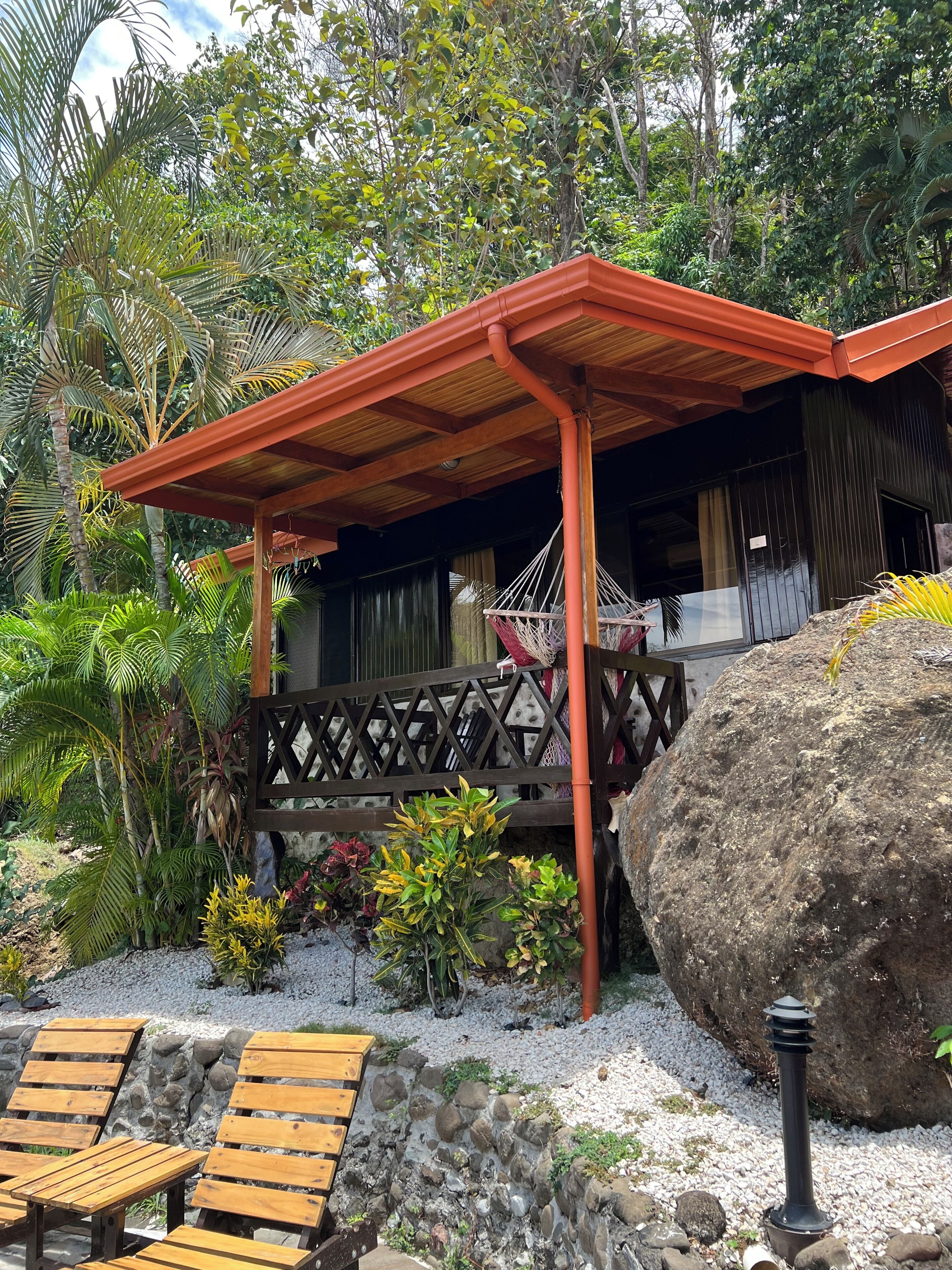 A covered outdoor area with a wooden bench, a hammock, and a large rock.