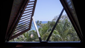 Une vue à travers une fenêtre à persiennes en bois, donnant sur un paysage tropical avec des palmiers et un ciel bleu azur.