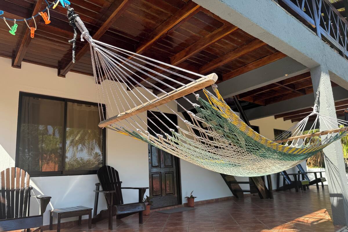 A hammock hanging between wooden beams in a covered outdoor area with tiled flooring, chairs, and a door.