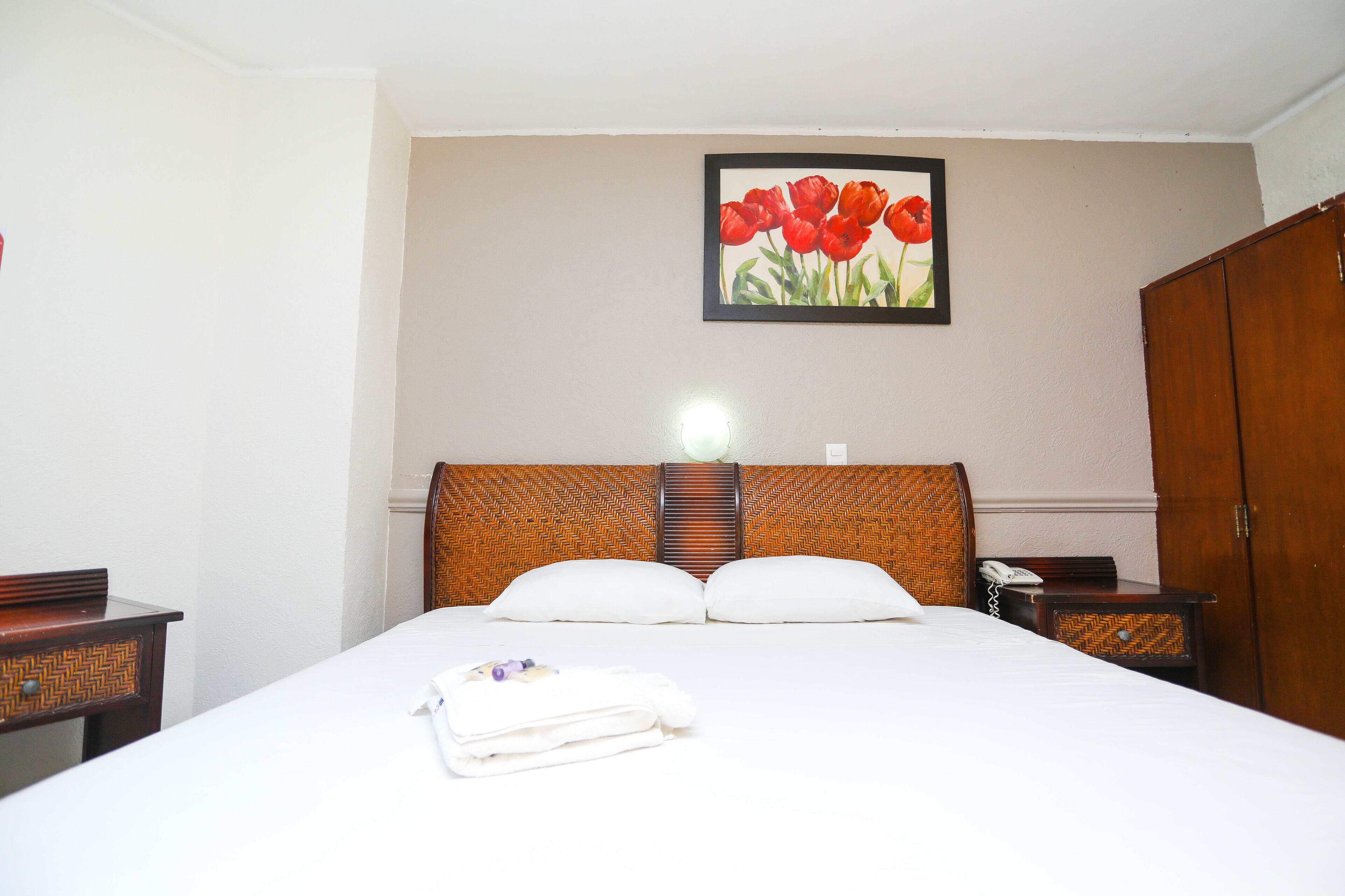 A hotel room with a bed, a headboard, a bedside table with a lamp, and a painting of red flowers on the wall.