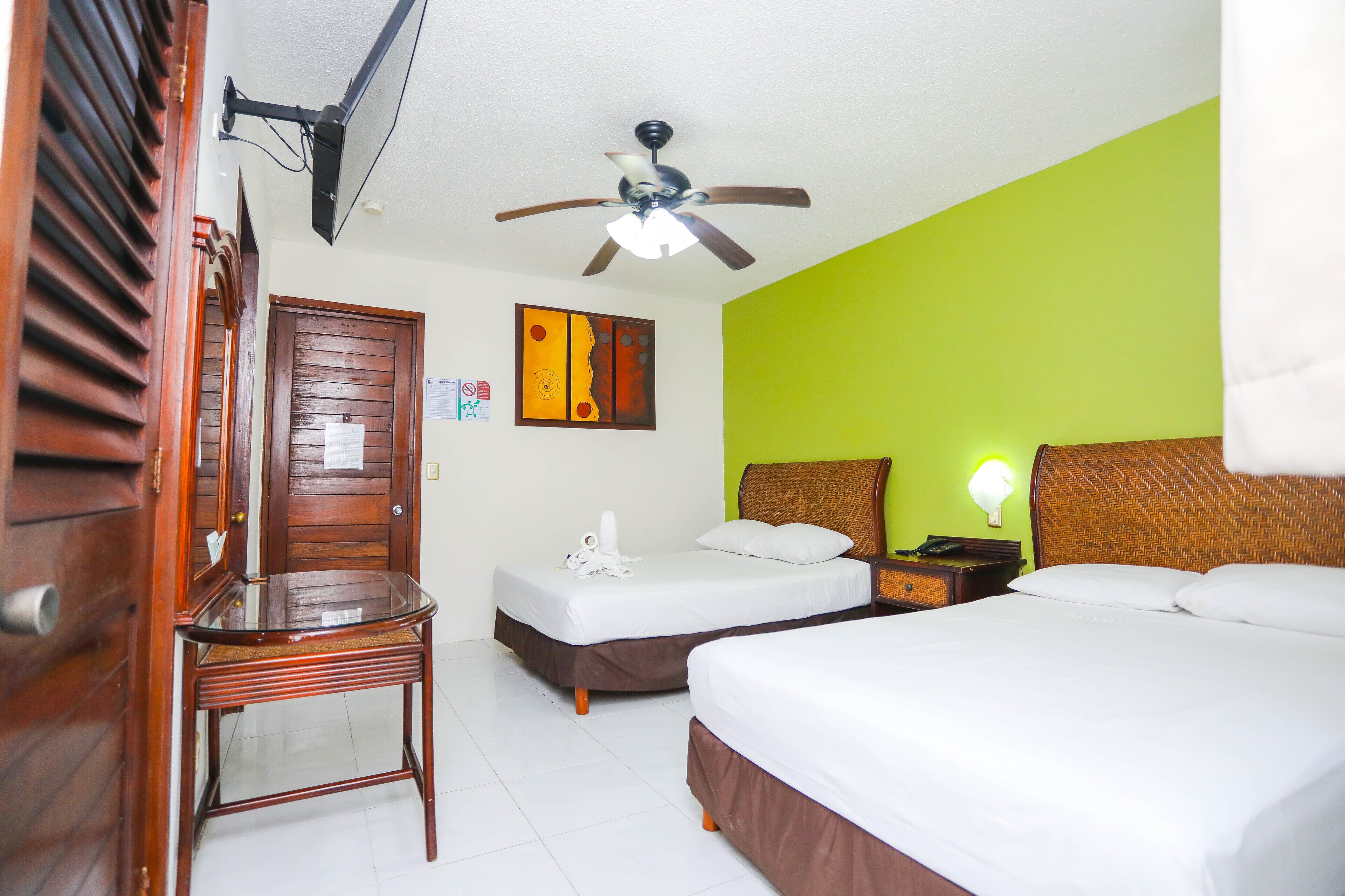 Une chambre d’hôtel avec deux lits, un ventilateur de plafond, une porte en bois, une table en verre et un mur vert.