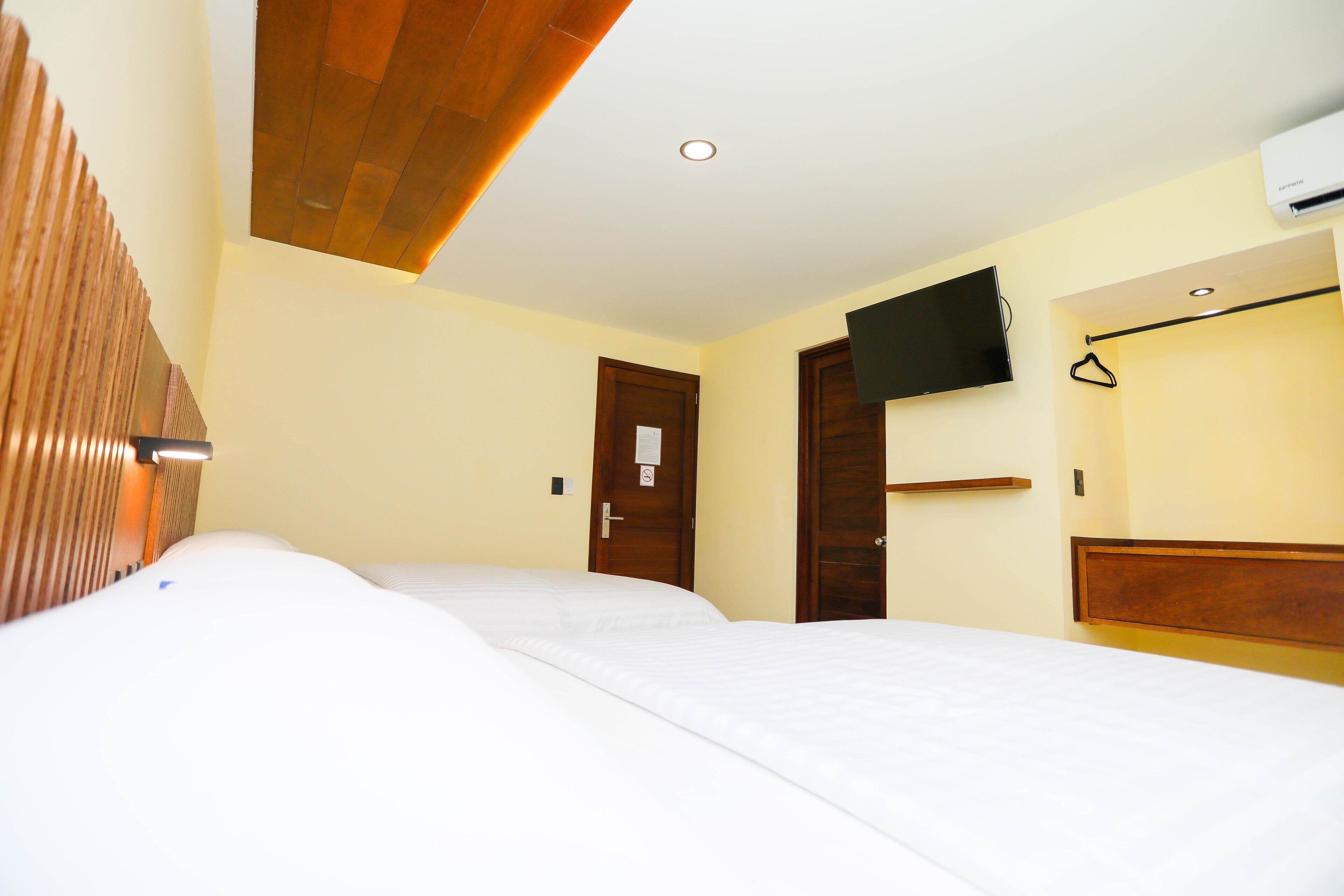A hotel room with a wooden headboard, a flat-screen TV mounted on the wall, and a bed with white bedding.