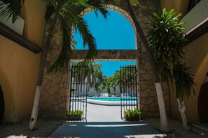 Une entrée menant à un espace piscine, agrémentée d’une arche en pierre, de palmiers et d’un ciel d’un bleu limpide.