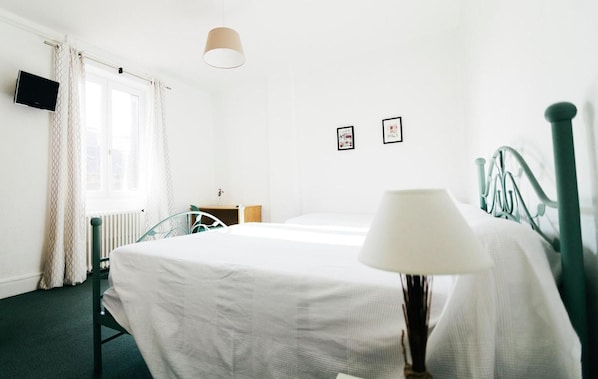 A hotel room with a bed, a bedside table, a lamp, a radiator, a TV, and a window with curtains. - Hôtel Restaurant Le Lion dOr (Meximieux)