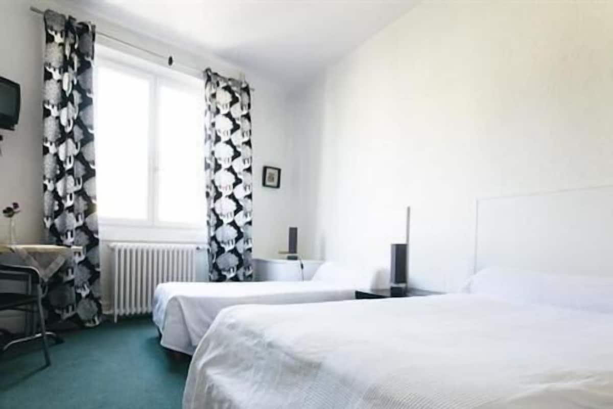 Habitación de hotel con dos camas, un escritorio, una silla, un radiador y una ventana con cortinas.
