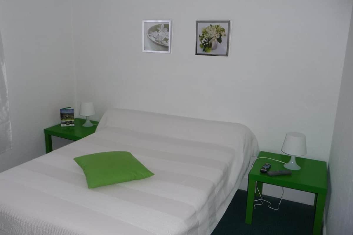 Habitación de hotel ordenada con una cama individual, un almohadón verde, dos mesitas de noche con lámparas y dos cuadros en la pared.