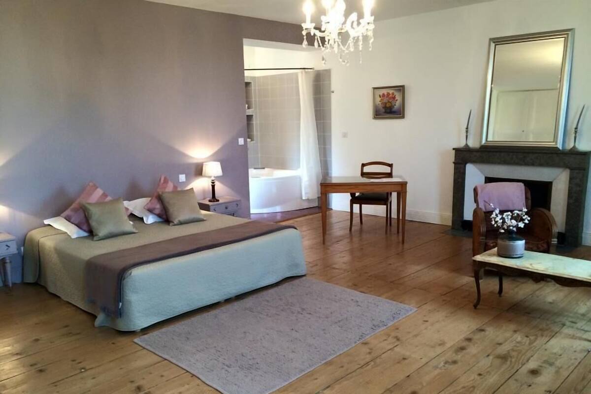 A bedroom with a bed, bedside tables, a chair, a fireplace, and a painting on the wall.