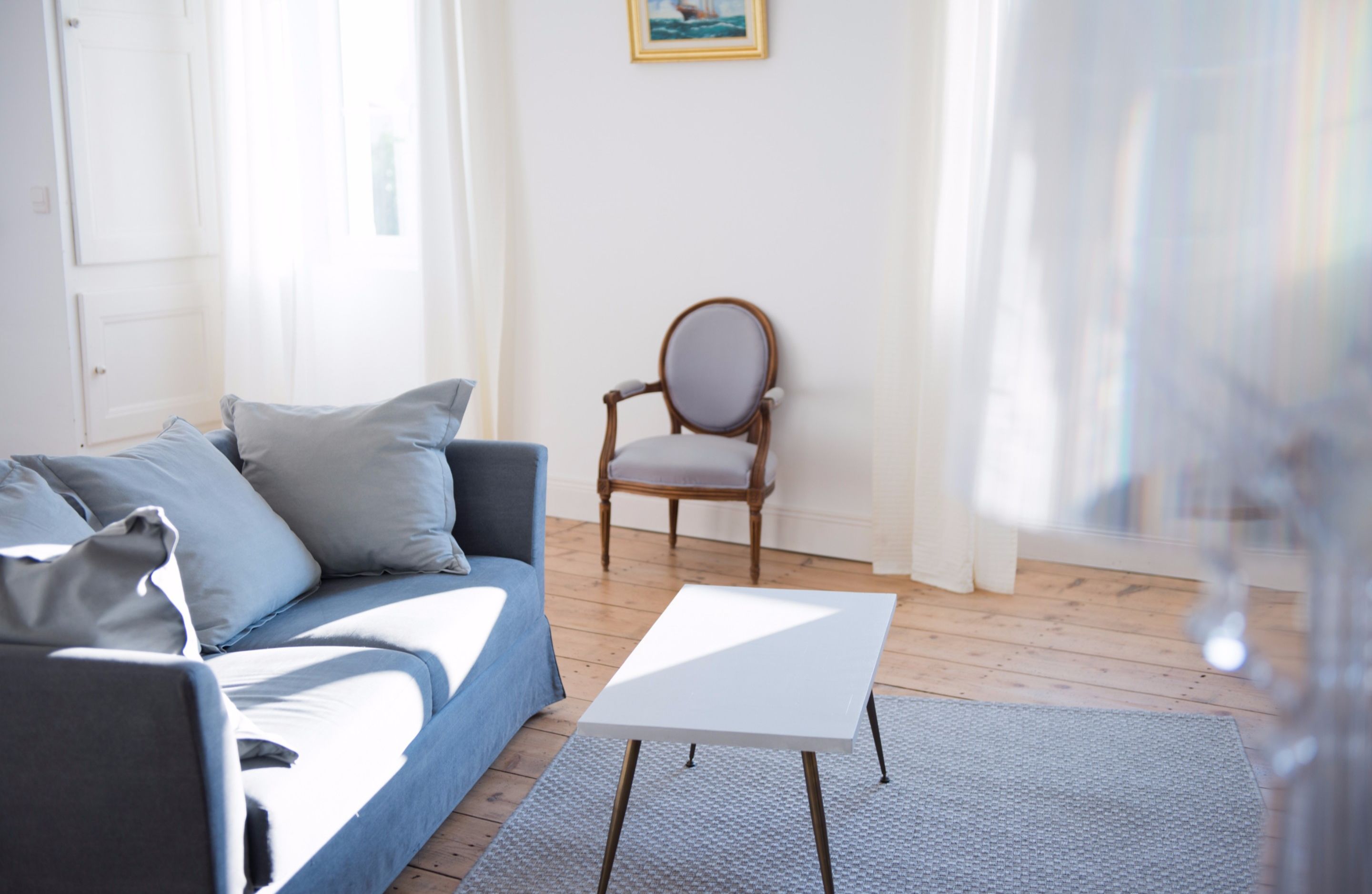 A living room with a blue sofa, a white coffee table, a wooden chair, and a painting on the wall.
