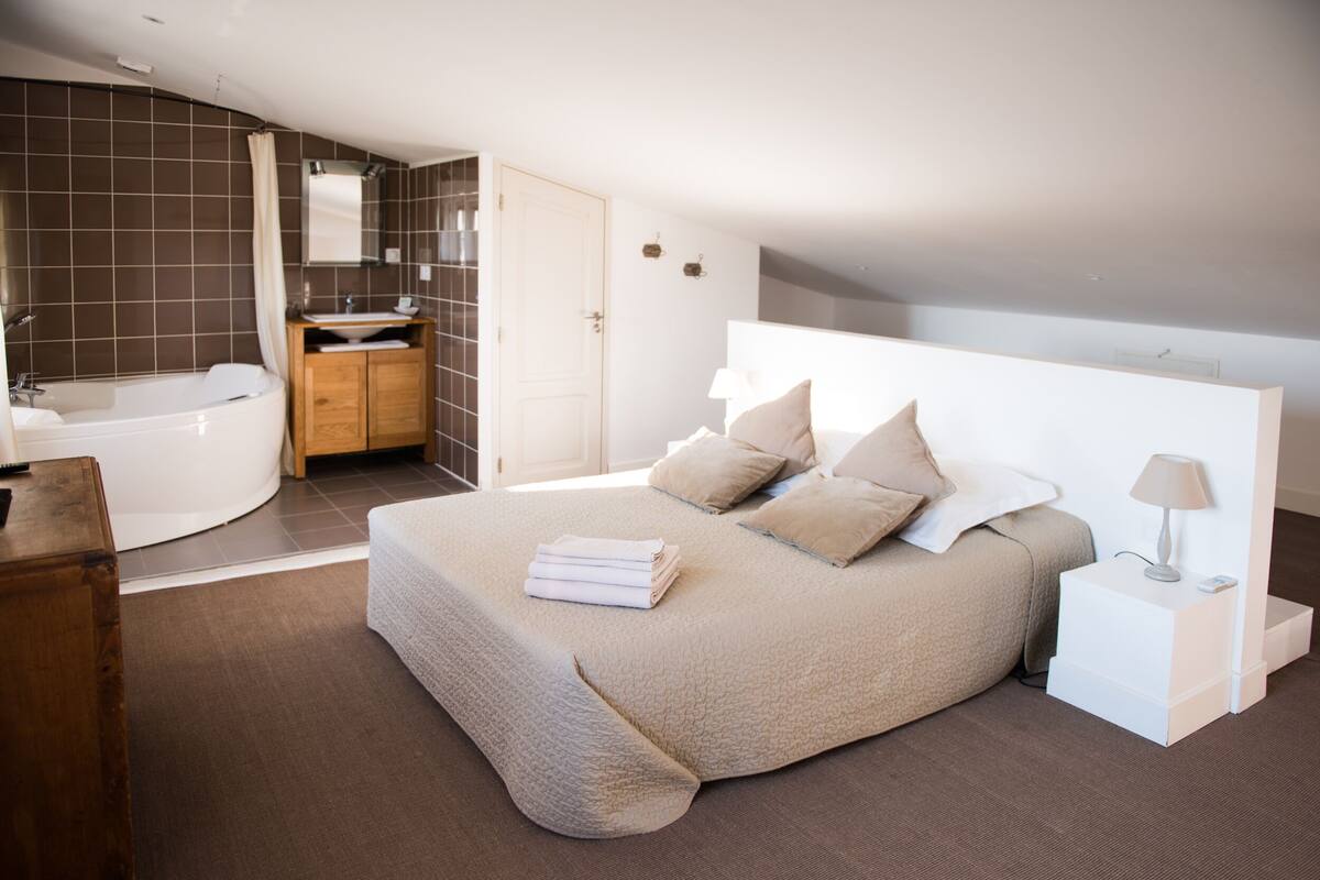 A bedroom with a large bed, a bedside table, a nightstand, a lamp, a bathroom with a bathtub, and a wooden cabinet.