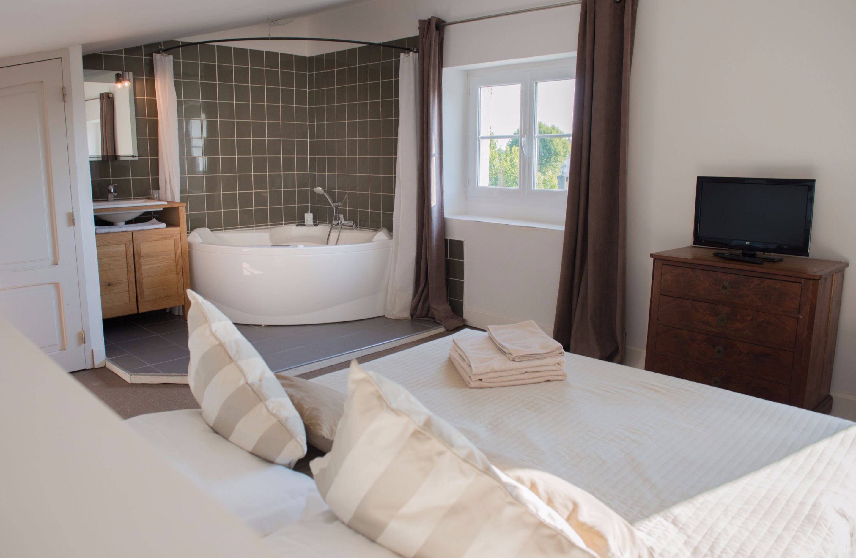 A bedroom with a large bed, a TV on a dresser, a bathroom with a bathtub, and a window with curtains.