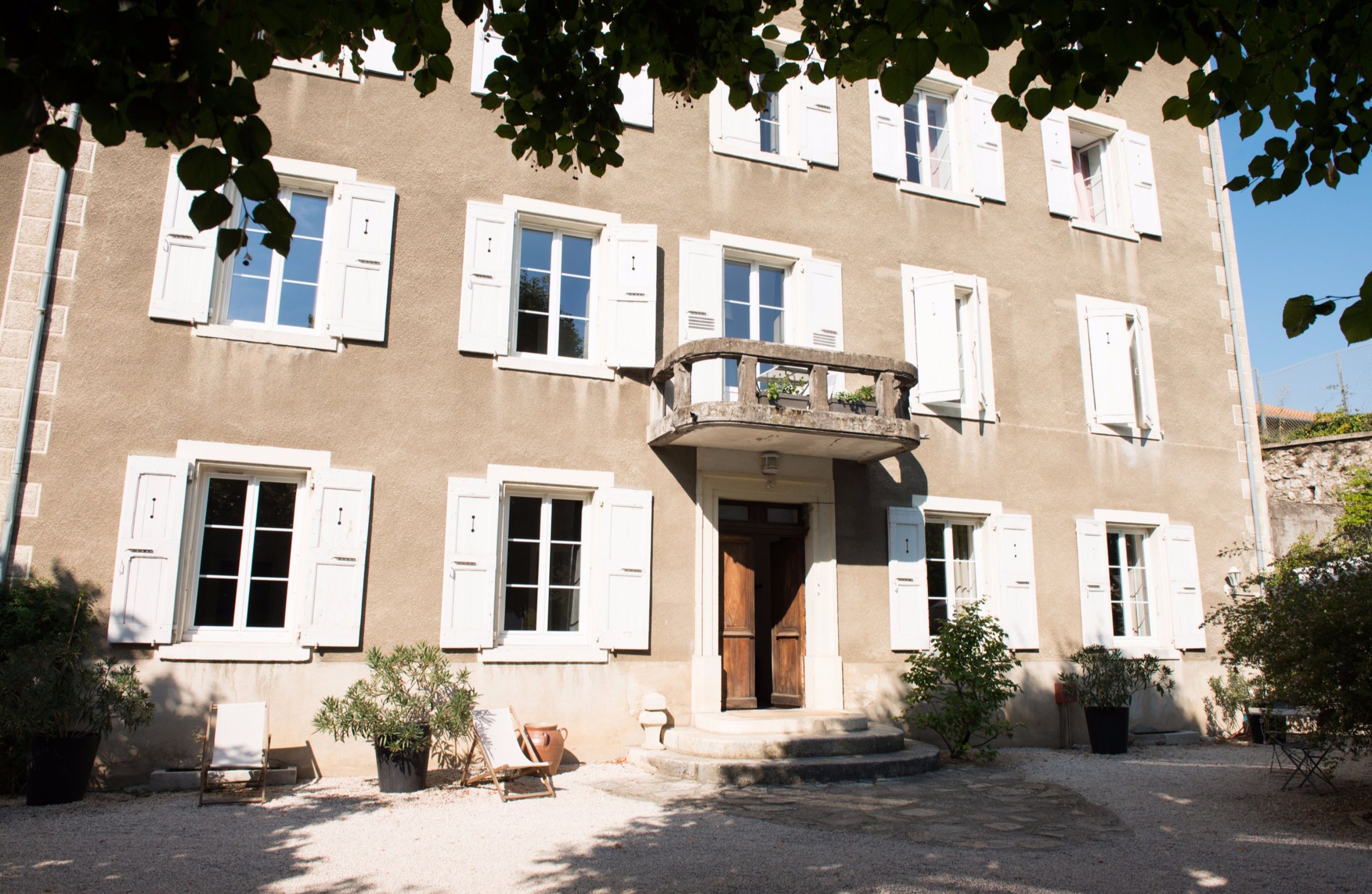 A two-story building with white shutters, a balcony, and a gravel courtyard with potted plants.