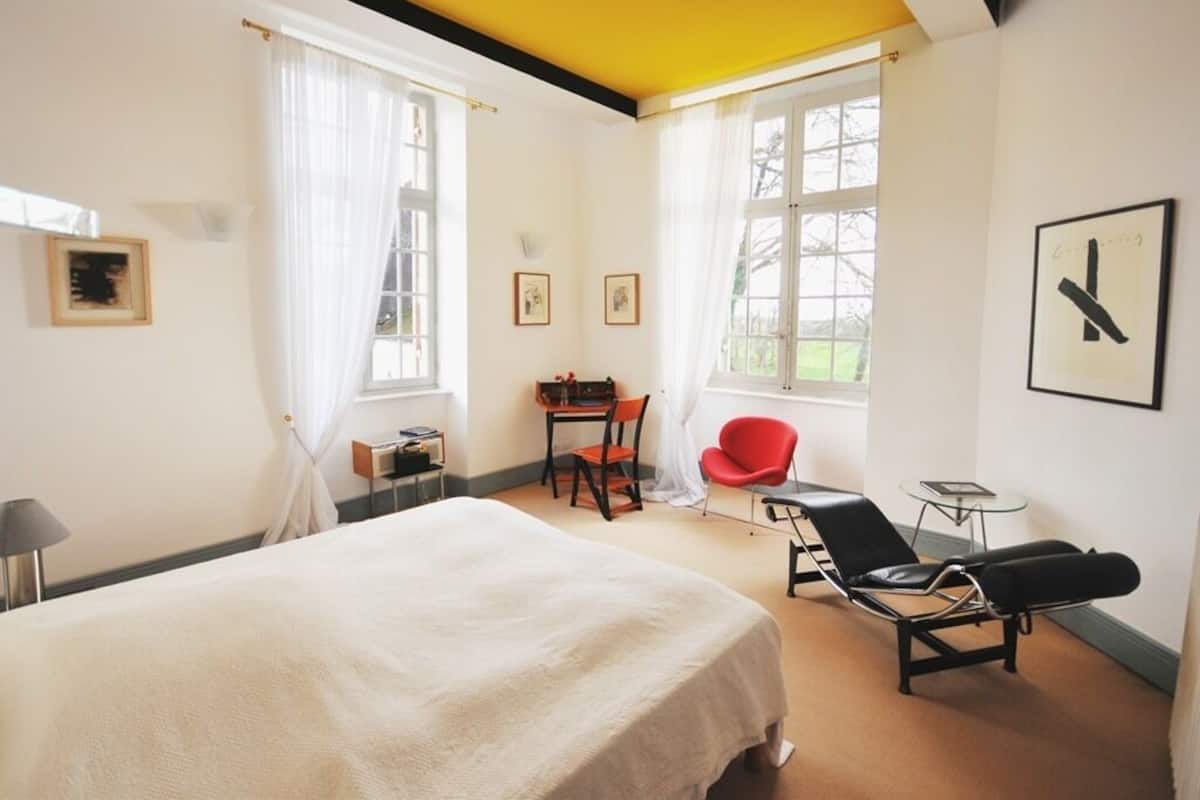 Ein Hotelzimmer mit Bett, Schreibtisch, einem roten Sessel, einem schwarzen Liegestuhl und einem Fenster mit Vorhängen.