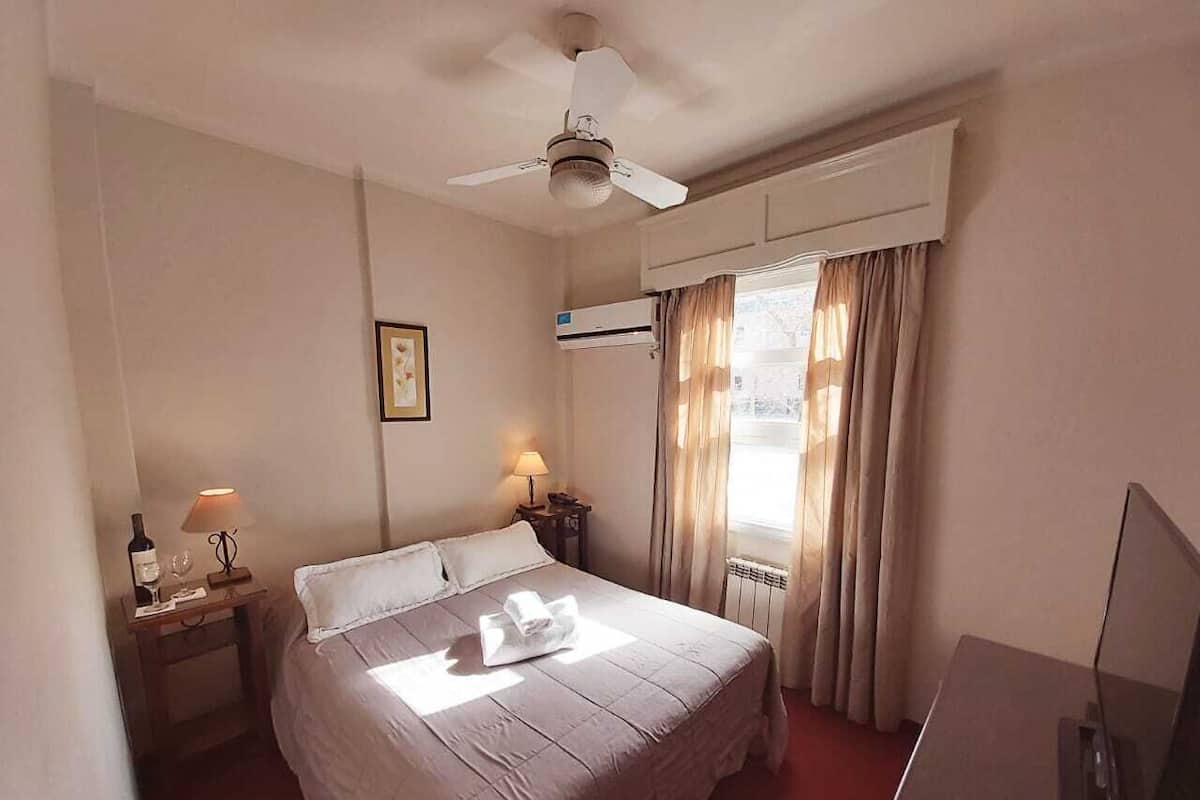 A hotel room with a bed, a desk, a television, a ceiling fan, and a window with curtains.