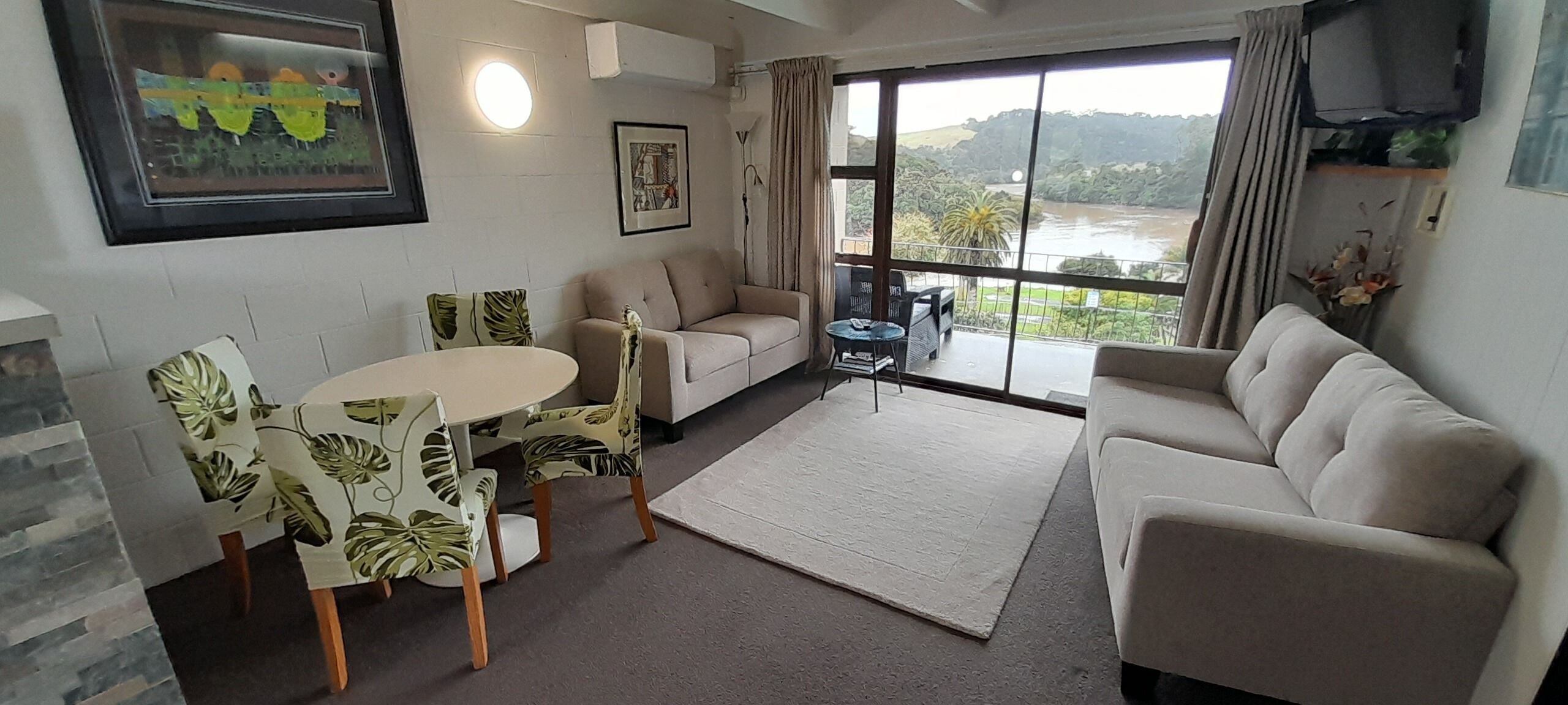 Un salon comprenant un canapé, une table à manger avec des chaises, un téléviseur fixé au mur et une grande fenêtre donnant sur une rivière et des collines.