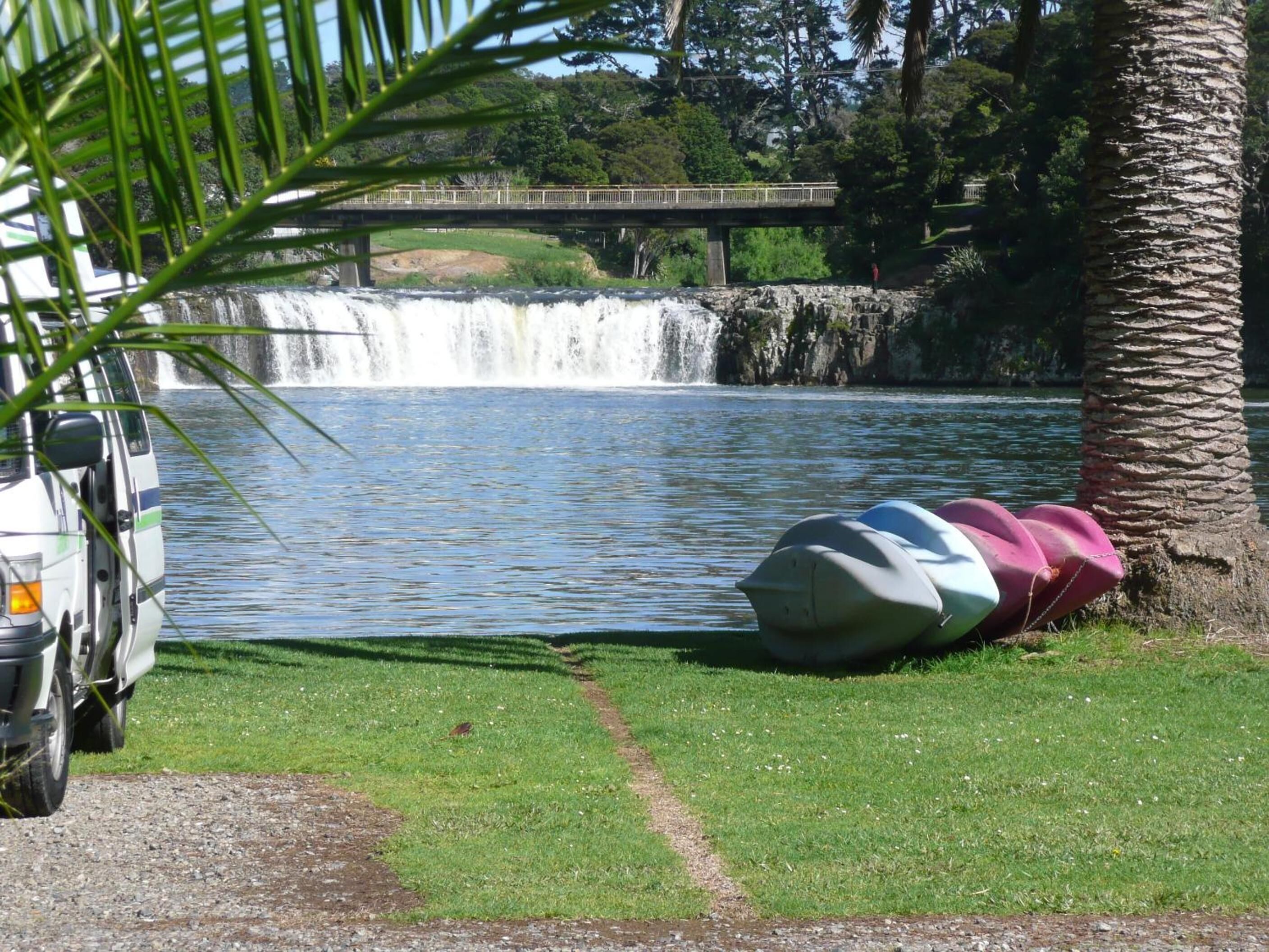 Un vehículo estacionado junto a un cuerpo de agua, con una cascada al fondo y un puente sobre el agua.