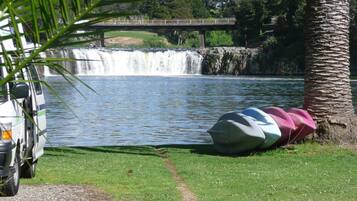 Un vehículo estacionado junto a un cuerpo de agua, con una cascada al fondo y un puente sobre el agua.