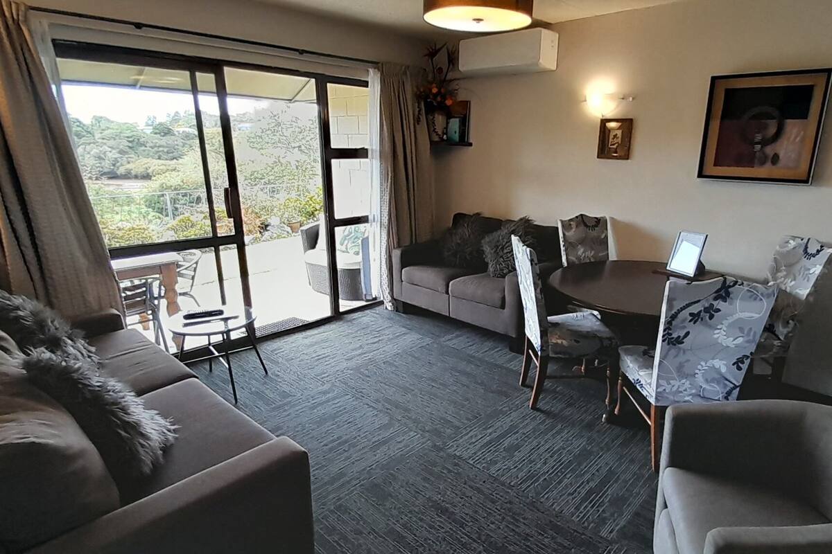 A room with a sofa, a dining table with chairs, a balcony with a view of greenery, and a wall-mounted air conditioner.