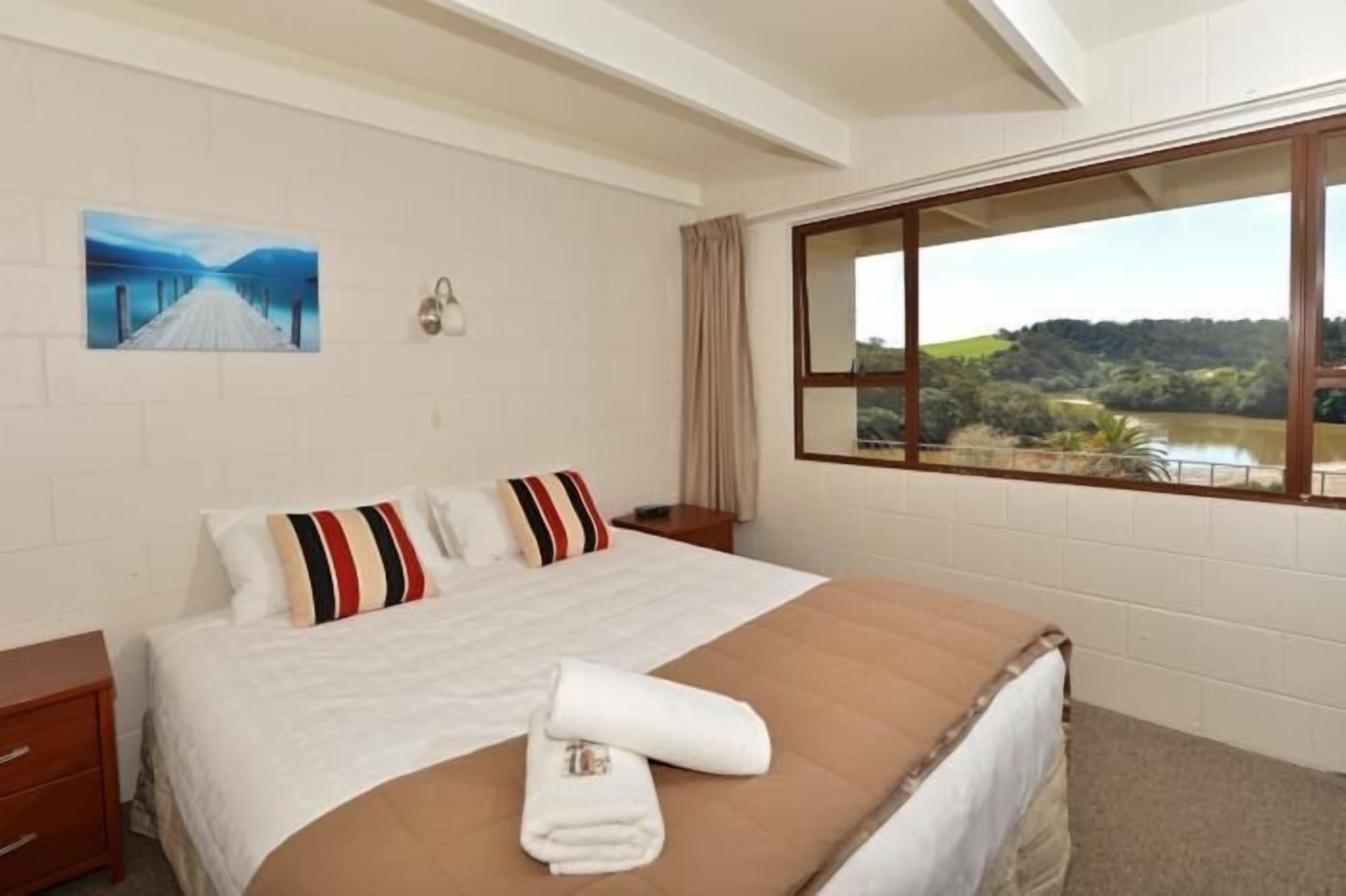 A hotel room with a large bed, a painting on the wall, a window with a view of greenery, and a nightstand with a lamp.