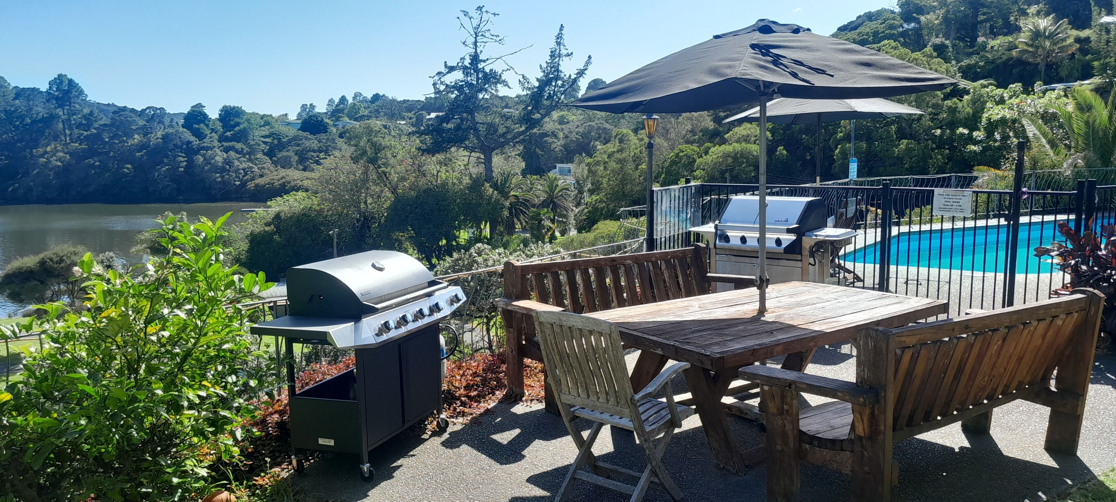 A patio with a grill, wooden table, and chairs, overlooking a lake and forested hills.