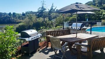 A patio with a grill, wooden table, and chairs, overlooking a lake and forested hills.