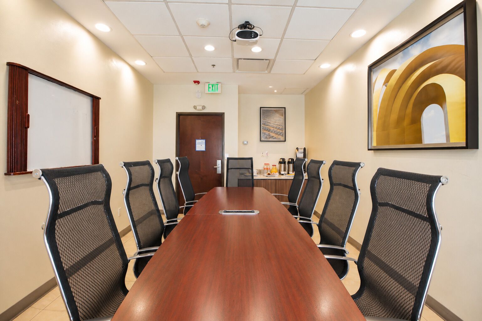 A conference room with a long wooden table, six mesh chairs, a projector, an exit sign, and a framed artwork on the wall.