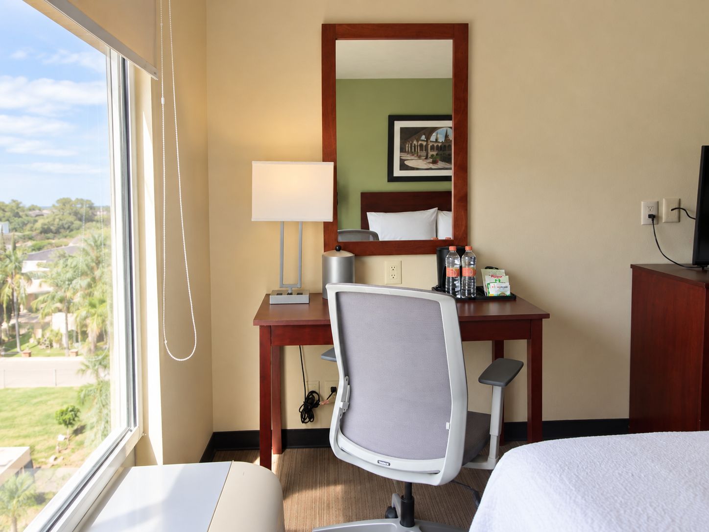 Een hotelkamer met een bed, een bureau met een stoel, een lamp, een spiegel, een televisie en een raam met uitzicht op groen.