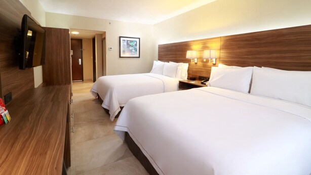 Una habitación de hotel con dos camas, un televisor montado en la pared, una mesita de noche con lámpara y un cuadro enmarcado en la pared.
