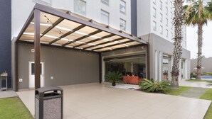 Entrada de hotel moderna con un porche cubierto, un contenedor de basura y una palmera.