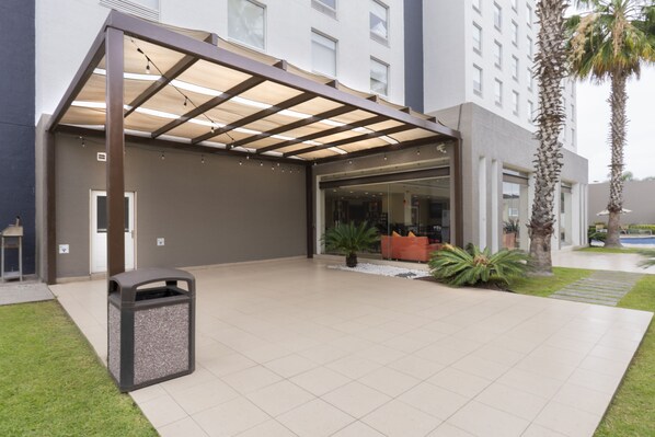 A modern hotel entrance with a covered patio, a trash bin, and a palm tree. - Holiday Inn Express Guadalajara Aeropuerto by IHG (El Salto)