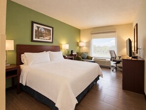Ein Hotelzimmer mit einem großen Bett, einem Schreibtisch mit Computer, einem Sessel, einem Fenster mit Aussicht und einem gerahmten Bild an der Wand.