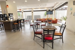 Un amplio comedor interior con varias mesas y sillas, una estación de café y una zona de estar con sofás y una pantalla de televisión.