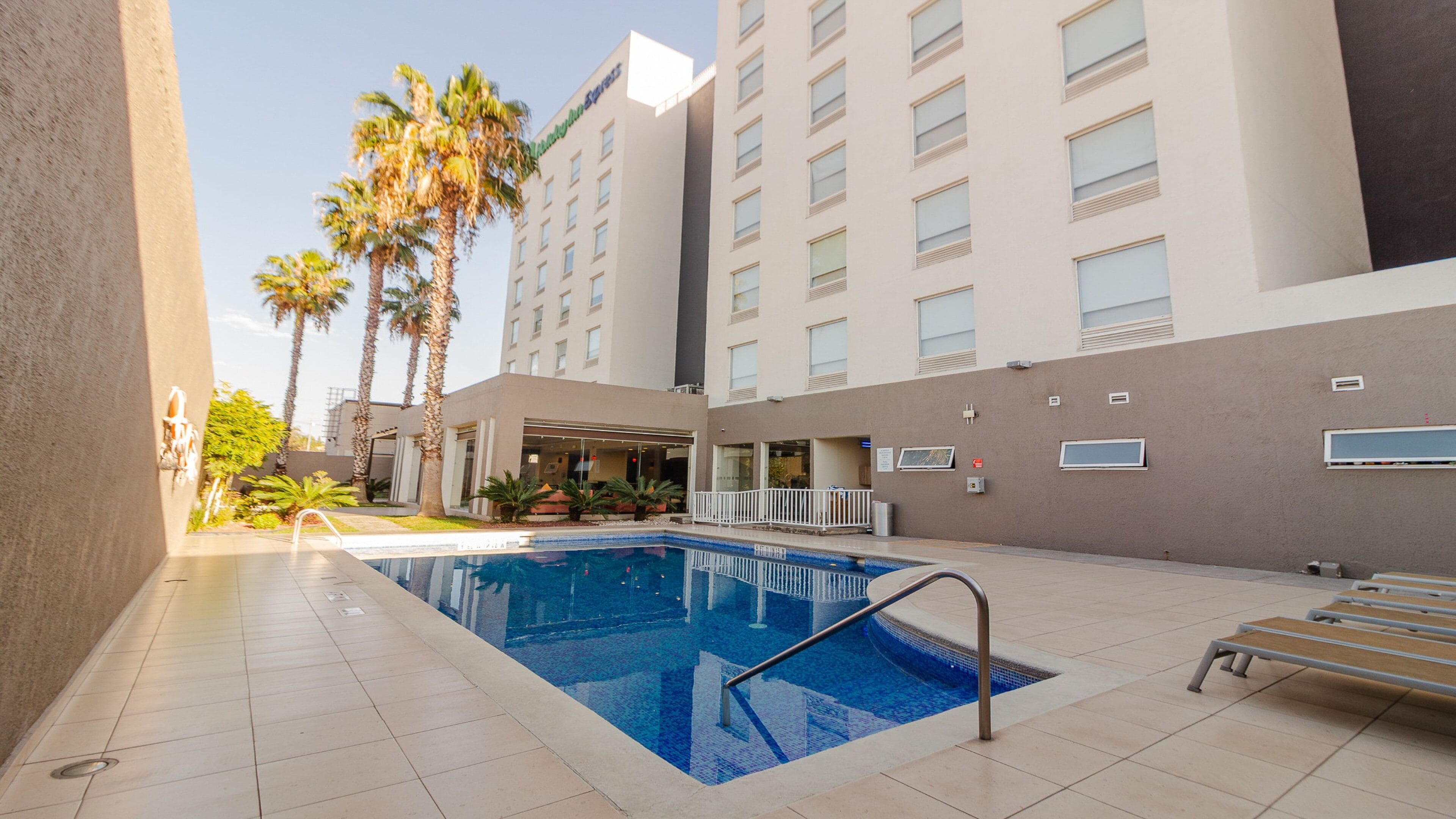 Una piscina con borde de baldosas, rodeada de palmeras y un edificio con varias ventanas.