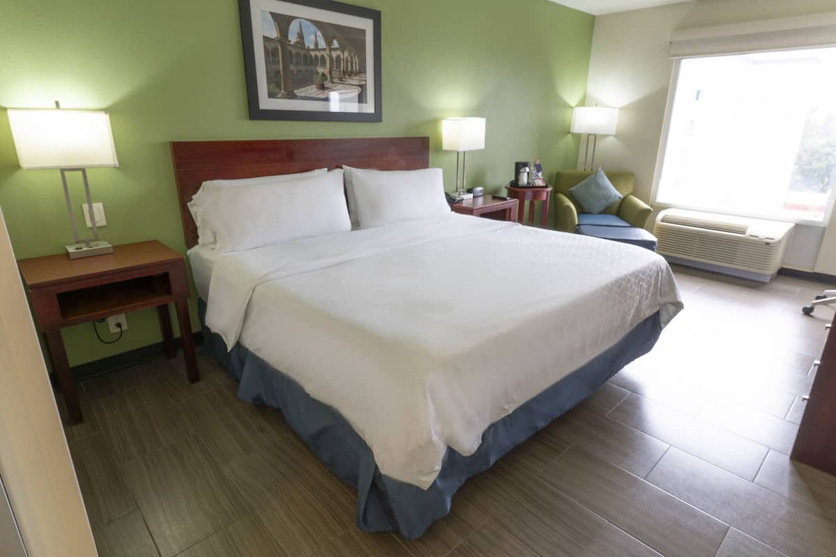 Une chambre d’hôtel avec un grand lit, deux tables de chevet, une petite table, un bureau, une chaise, une fenêtre avec vue et un tableau accroché au mur.
