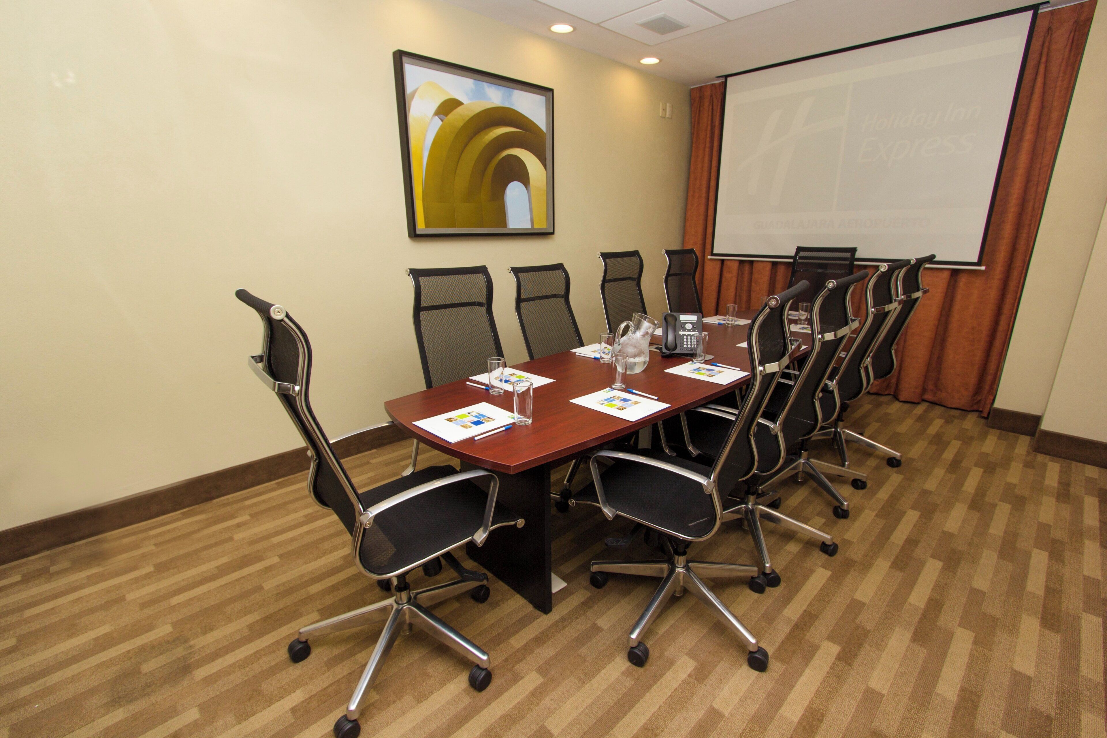 A conference room with a long table, chairs, a projector screen, and a painting on the wall.