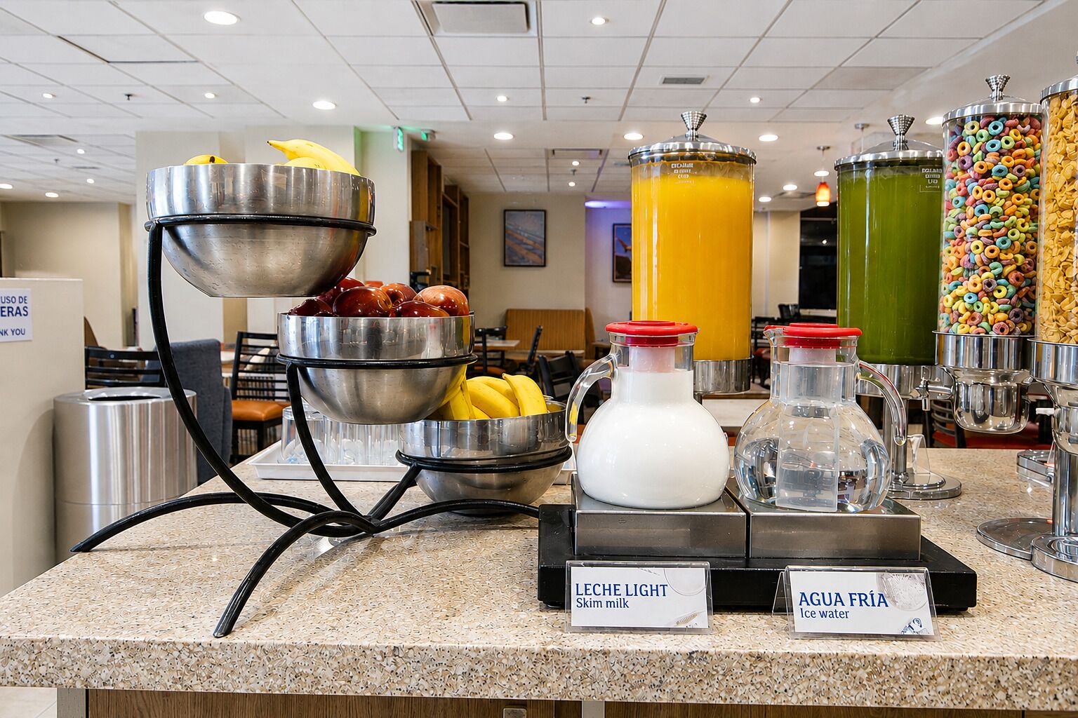 Mostrador de buffet con diversas opciones de comida y bebida, incluyendo un tazón de frutas, una jarra de leche y recipientes de agua y jugo.