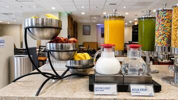 Mostrador de buffet con diversas opciones de comida y bebida, incluyendo un tazón de frutas, una jarra de leche y recipientes de agua y jugo.