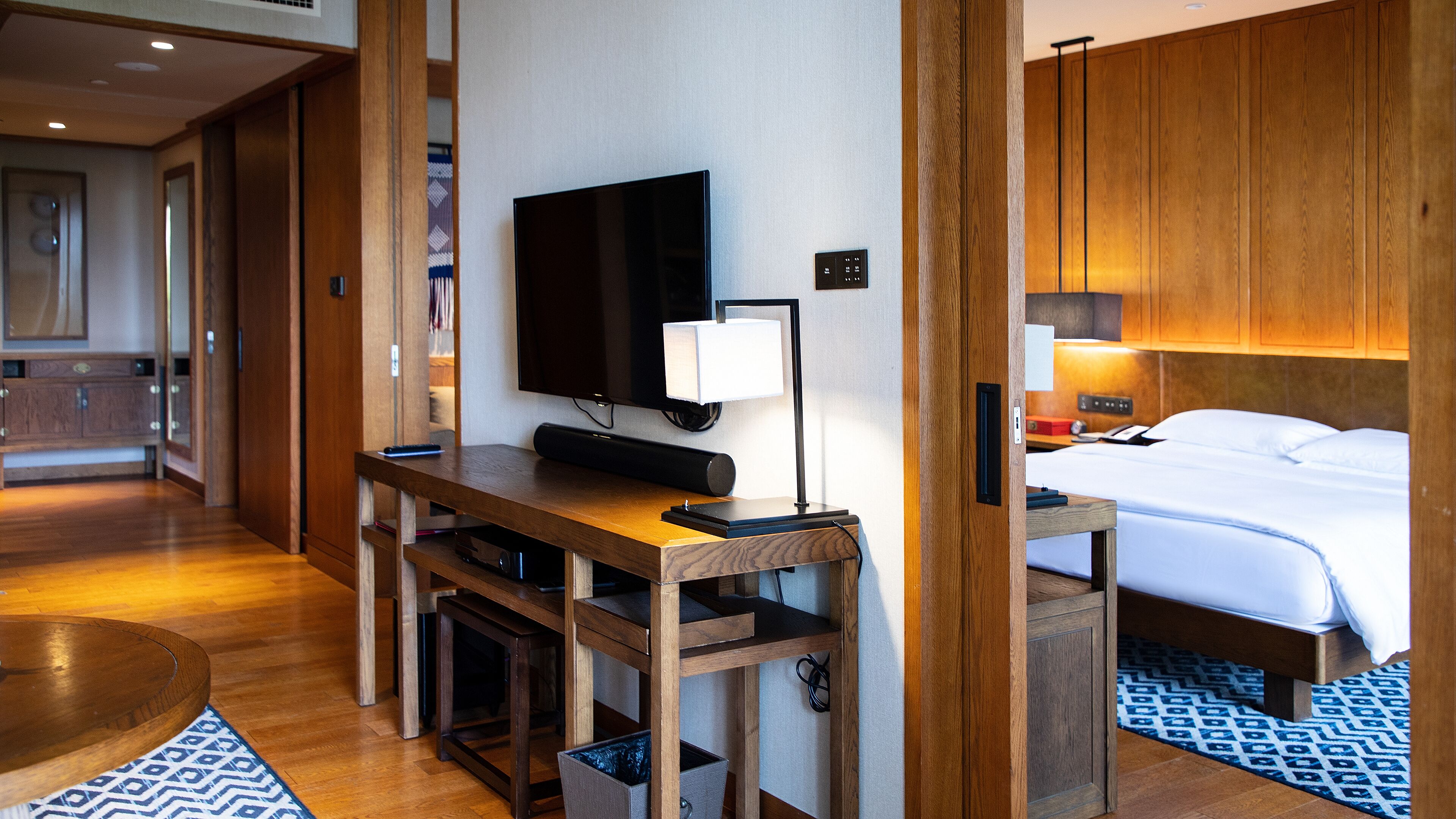 A hotel room with a wooden desk, a flat-screen TV, a bedside table with a lamp, a bed with white linens, and a patterned rug.