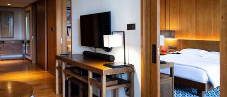 A hotel room with a wooden desk, a flat-screen TV, a bedside table with a lamp, a bed with white linens, and a patterned rug.