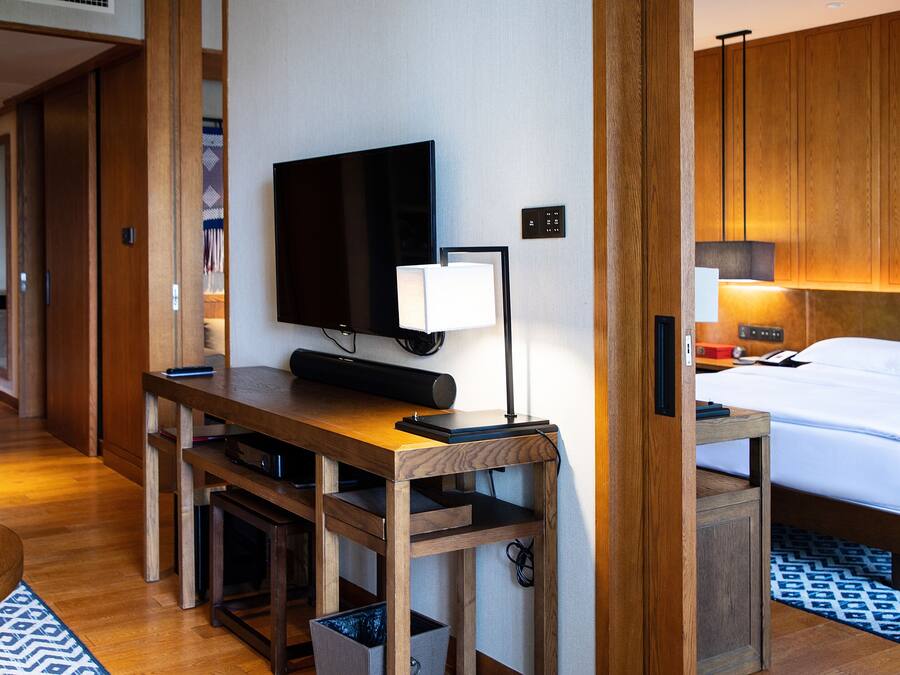 A hotel room with a wooden desk, a flat-screen TV, a bedside table with a lamp, a bed with white linens, and a patterned rug.