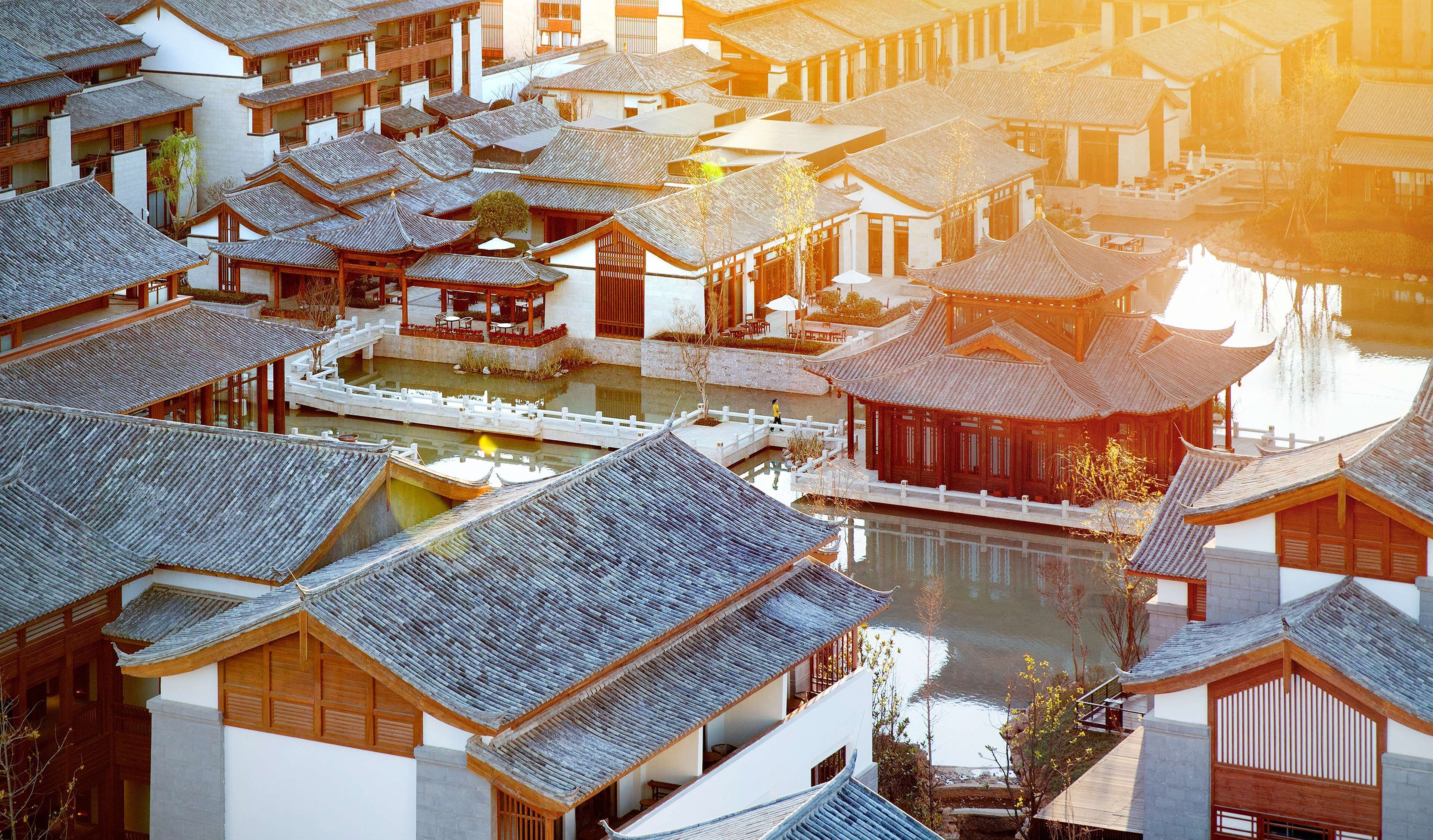 A traditional Chinese courtyard with multiple buildings, a central water feature, and a wooden bridge.
