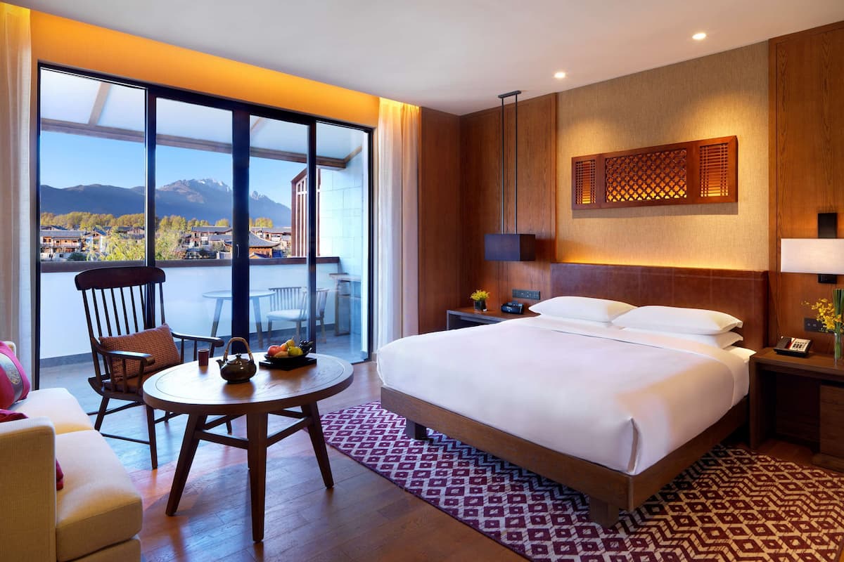 Ein Hotelzimmer mit einem großen Bett, einem Schreibtisch mit Stuhl, einem kleinen Tisch mit Obst, einem Nachttisch mit Lampe und Blick auf Berge durch große Fenster.