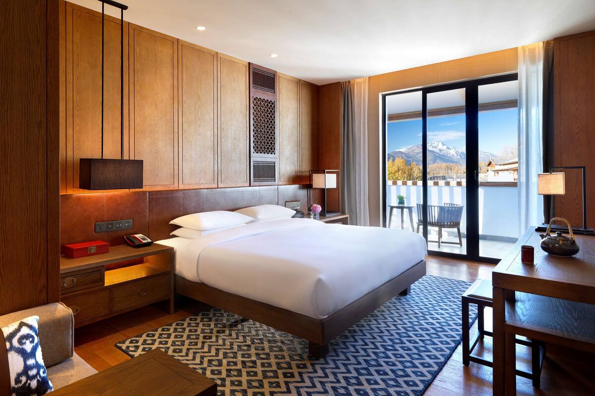 Ein Hotelzimmer mit einem großen Bett, Holzwandverkleidung, einem Schreibtisch mit Lampe, einem Sessel und Blick auf Berge durch das Fenster.