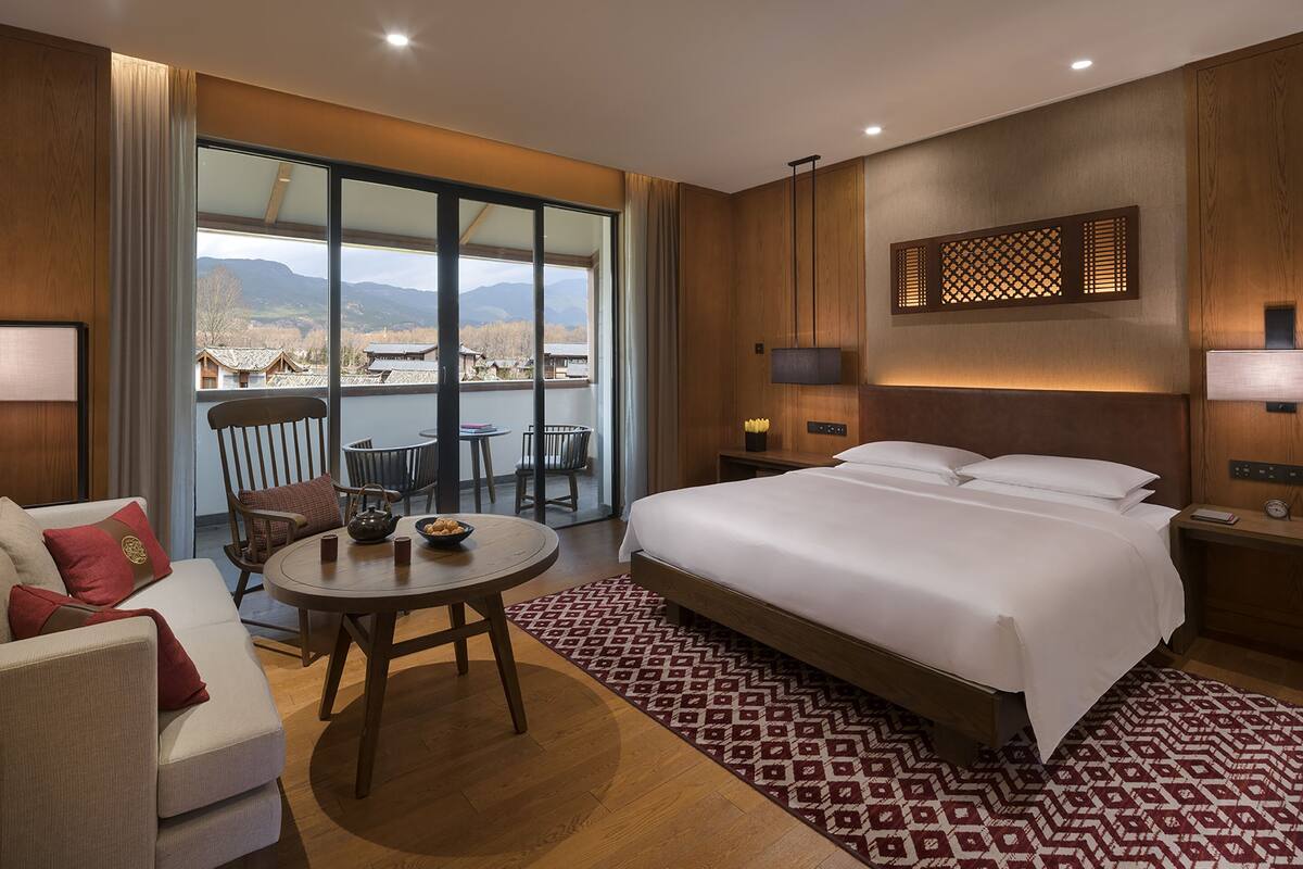 Ein Hotelzimmer mit einem großen Bett, einem Esstisch, einer Couch und Blick auf Berge durch ein großes Fenster.