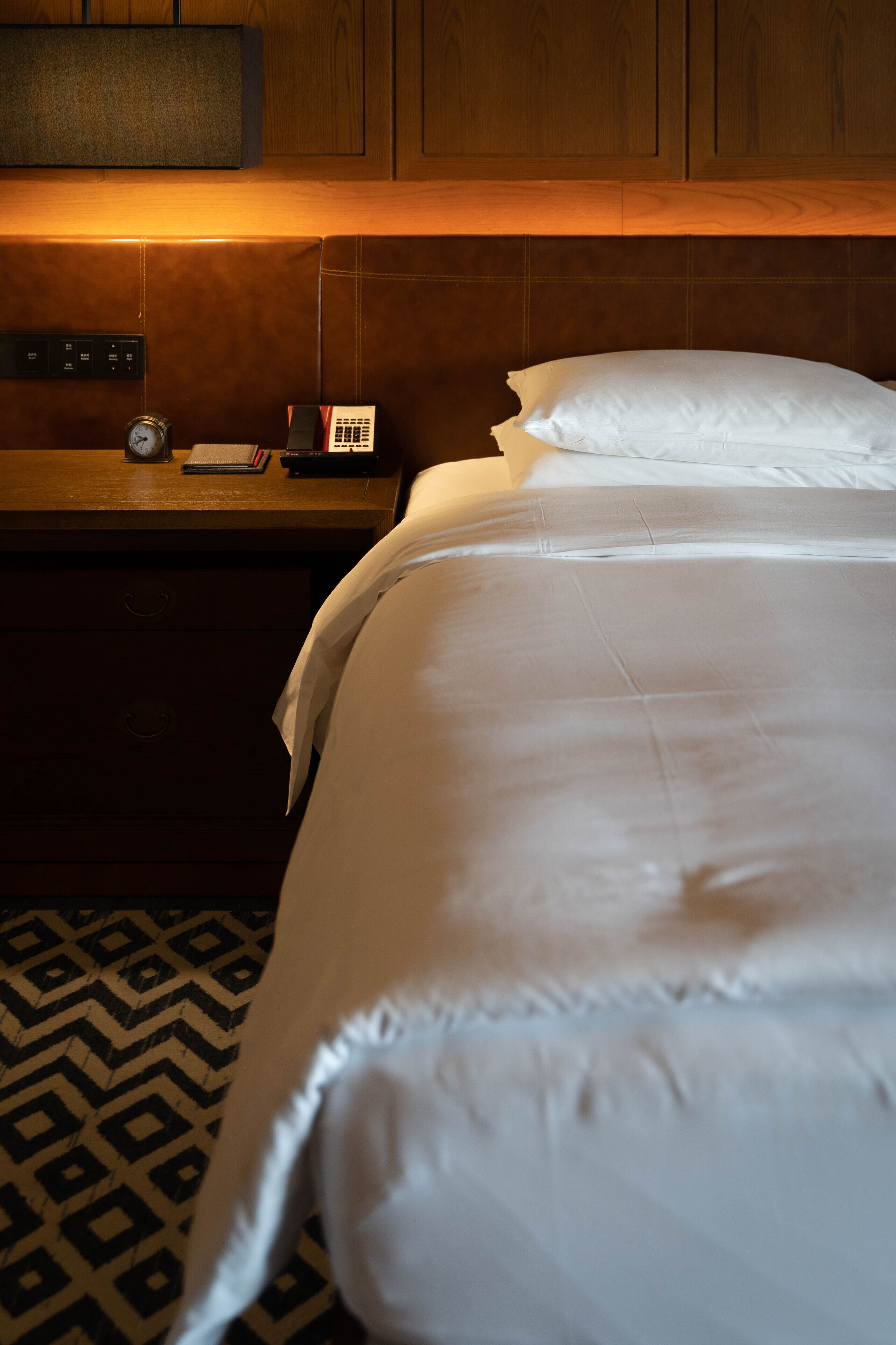 A hotel room with a bed, a bedside table with a clock, a remote control, and a patterned rug.