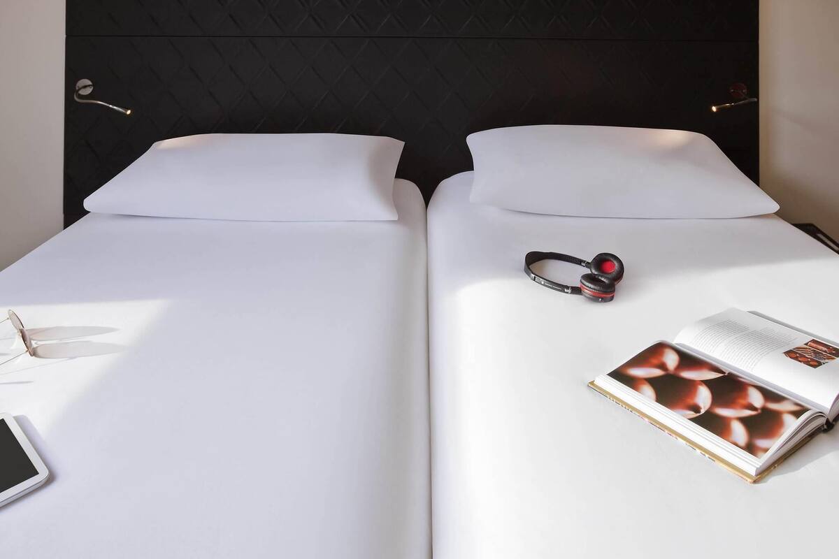 Two beds with white linens, a pair of headphones, a smartphone, and a book on one bed.