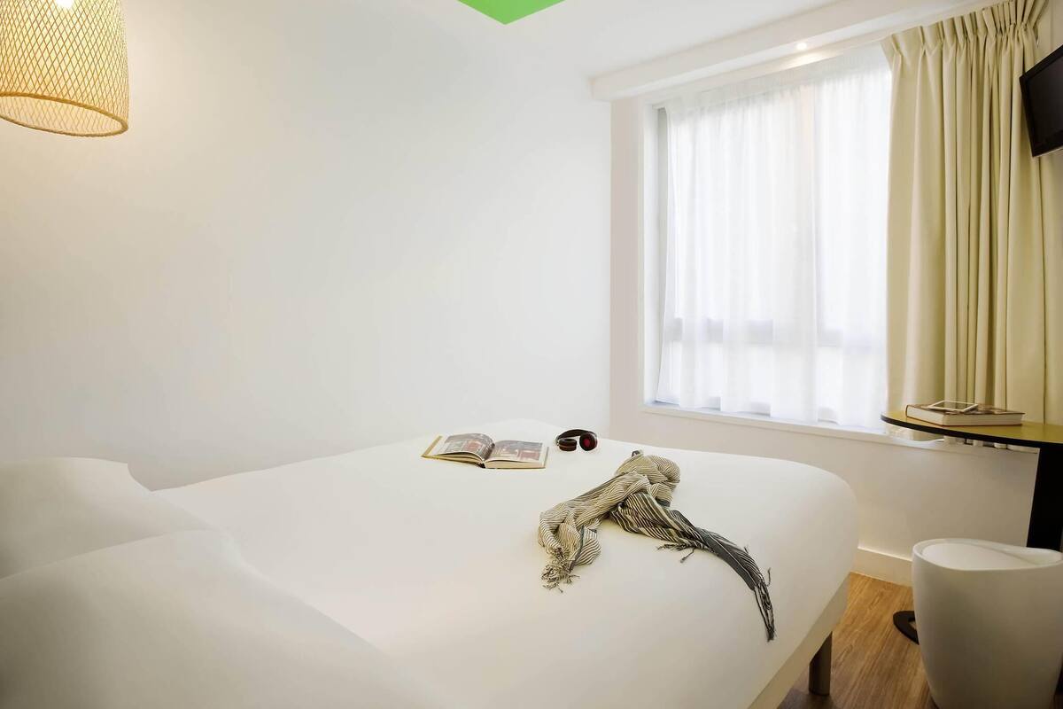 A hotel room with a bed, a window with curtains, a wall-mounted TV, and a hanging light fixture.