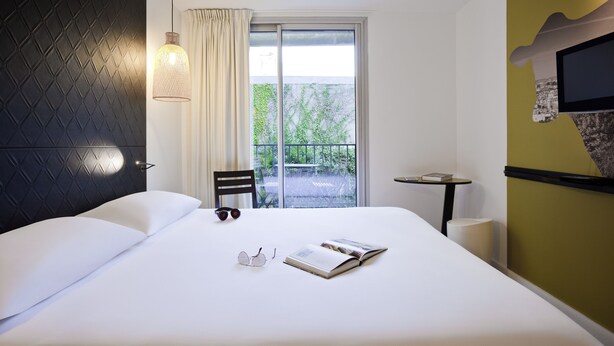 A hotel room with a bed, a chair, a small table, a TV, and a window with a view of greenery. - ibis Styles Paris Buttes Chaumont (Paris)