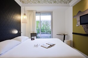 A hotel room with a bed, a chair, a small table, a TV, and a window with a view of greenery. - ibis Styles Paris Buttes Chaumont (Paris)