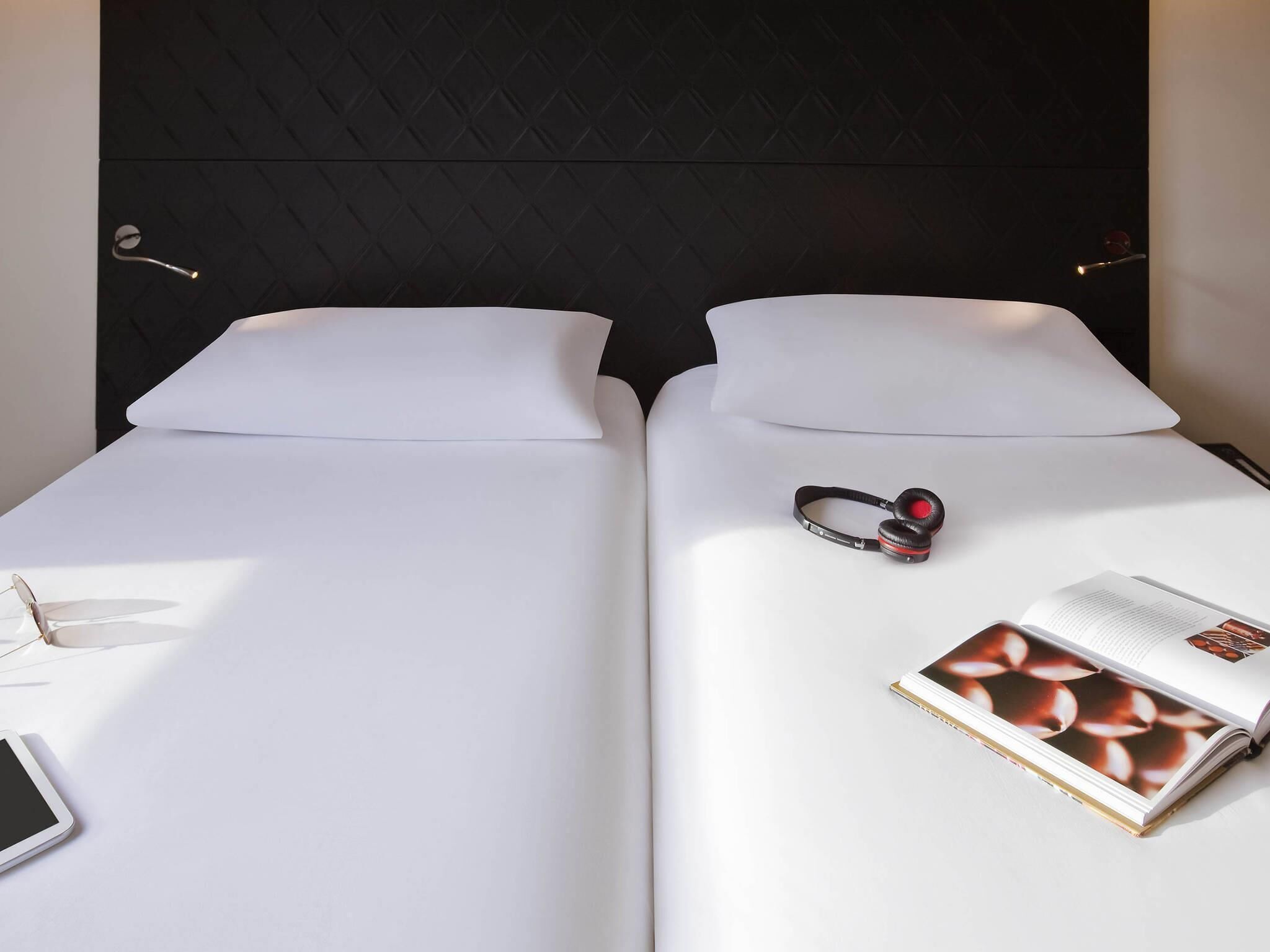 Duas camas com roupa de cama branca, um par de fones de ouvido, um smartphone e um livro sobre uma delas.