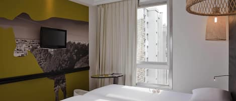 A hotel room with a bed, a mounted TV, a window with curtains, and a small table with a book and glasses.